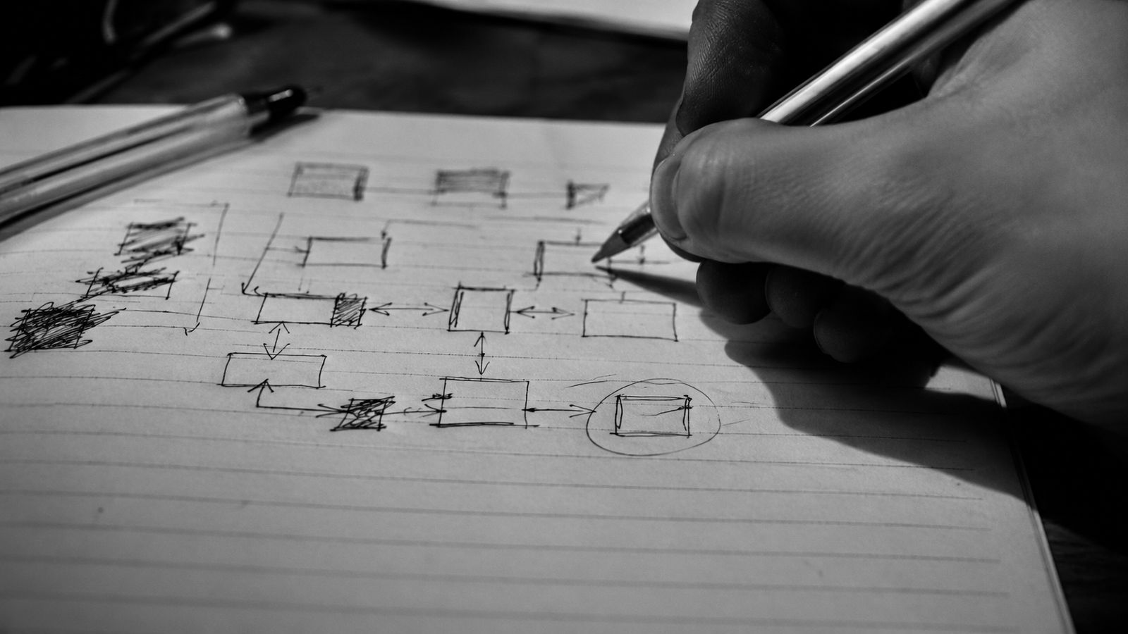 A hand drawing a flow diagram of boxes and arrows in ballpoint on a lined notebook.