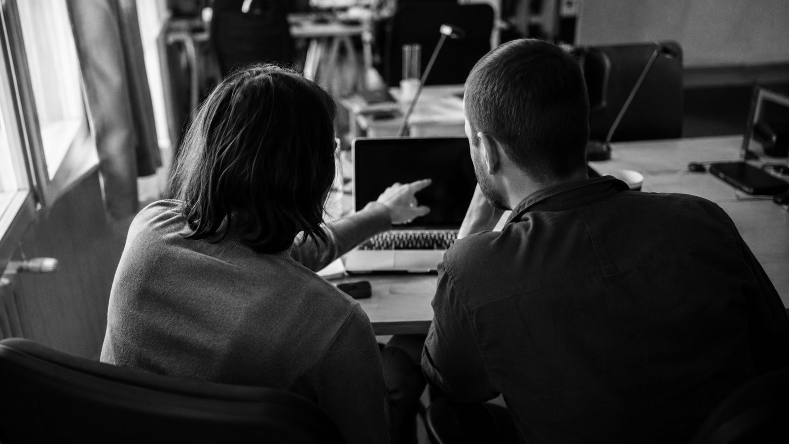 Two colleagues at one desk looking at the same laptop, one pointing at the screen.