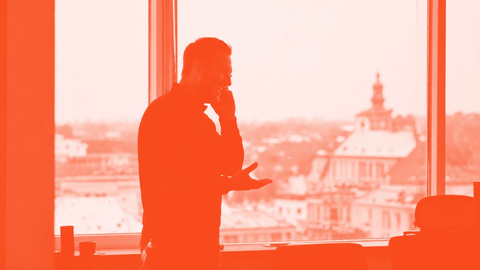 A person at an office window mid-conversation on the phone, backlit against the glass.